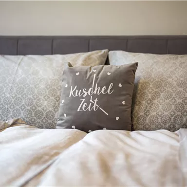 Ein bequemes Bett mit dekorativen Kissen und einer gemütlichen Decke. Auf einem Kissen steht der Schriftzug „Kuschel Zeit“.
