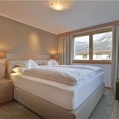 Ein modernes Schlafzimmer mit einem großen Doppelbett und einem Fenster, das Tageslicht hereinlässt. Die Wände sind in sanften Farbtönen gehalten, und es gibt eine kleine Nachttischlampe.