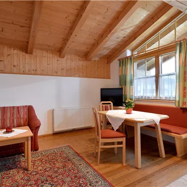 Ein gemütlicher Raum mit Holzwänden und -decke. Der Bereich enthält ein Sofa, einen Esstisch und große Fenster mit Vorhängen.