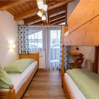 Ein gemütliches Schlafzimmer mit zwei Einzelbetten und einem Etagenbett. Helle Holzvertäfelung und Fenster mit Vorhängen sorgen für eine einladende Atmosphäre.