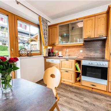 Eine gemütliche Küche mit Holzmöbeln und großen Fenstern. Auf dem Tisch stehen frische Rosen in einer Vase.