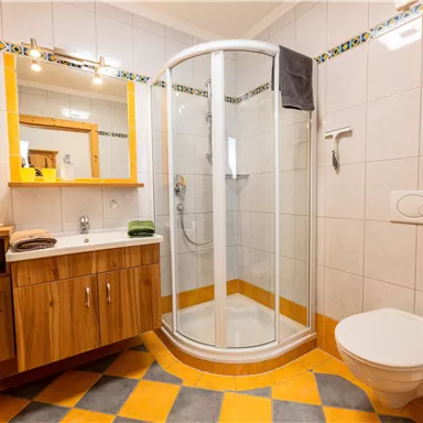 Ein helles Badezimmer mit einer Dusche, einem Waschbecken und einer Toilette. Die Wände sind gefliest und der Boden hat ein attraktives Muster aus gelben und grauen Fliesen.