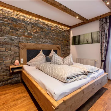 Ein gemütliches Schlafzimmer mit einem großen Holzbett und dekorativen Kissen. Die Wand aus Steinen und das warme Licht schaffen eine einladende Atmosphäre.