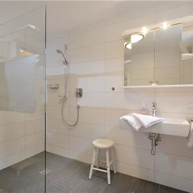 A modern bathroom with a glass shower and bright tiles. A beautiful wooden shelf with towels and a small stool complete the space.