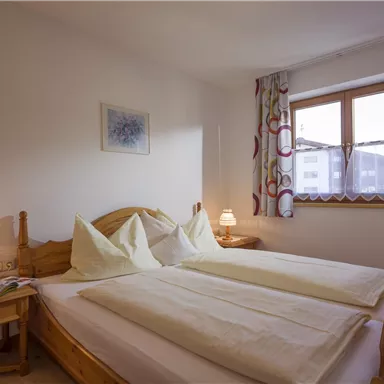 A cozy bedroom with two single beds and soft colors. Windows with drapes let in daylight, and a bedside lamp creates a pleasant atmosphere.