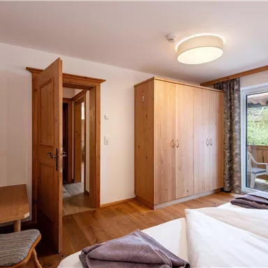 A bright bedroom with wooden furniture and a large window. There is a bed, a desk, and access to a balcony.