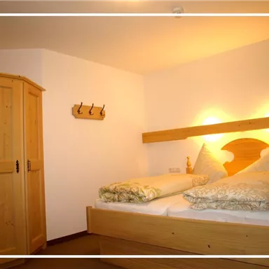 Ein gemütliches Schlafzimmer mit einem Doppelbett und dekorativen Kissen. Im Hintergrund sind ein Holzschrank und eine Garderobe zu sehen.