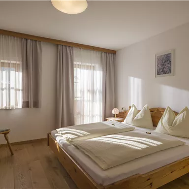 A bright bedroom with a double bed and large windows. The decor is simple and cozy, featuring a wooden chair and soft curtains.