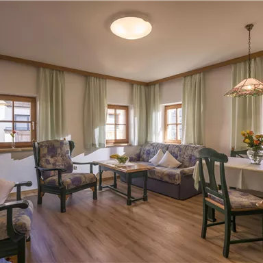 A cozy living room with comfortable furniture and large windows. The atmosphere is friendly and welcoming, with floral decor and soft colors.