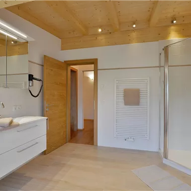 A modern bathroom with a wooden ceiling and elegant white furniture. It features a shower and plenty of light.