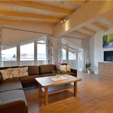 A modern living room with large windows and wooden floors. There is a cozy sofa, a table, and a television.