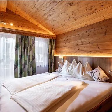 A cozy bedroom with a wooden ceiling and warm light. The bed is inviting, surrounded by windows and decorative curtains.