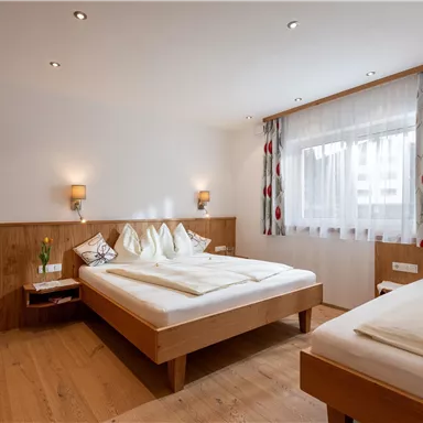 A cozy bedroom with two single beds and warm wooden interiors. The room features large windows with plenty of daylight and modern lighting.