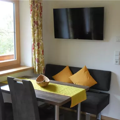 Ein gemütlicher Essbereich mit einem Tisch, zwei Stühlen und einer Bank. An der Wand hängt ein Fernseher und die Fenster sind mit bunten Vorhängen dekoriert.