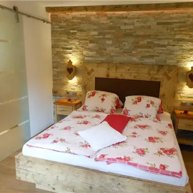 Ein gemütliches Schlafzimmer mit einem Doppelbett und floralem Bettzeug. Die Wände sind mit Stein dekoriert und es gibt warme Lichtquellen.