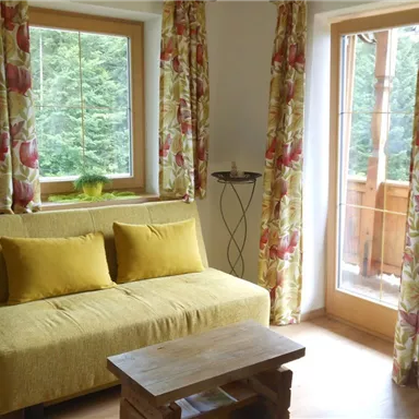 Ein helles Wohnzimmer mit einem gelben Sofa und einem Holzkaffee Tisch. Große Fenster bieten einen Blick auf die Natur.