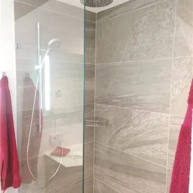 Eine moderne Dusche mit gläserner Kabine und grauen Fliesen. Rote Handtücher hängen an der Wand und verleihen dem Raum einen farbenfrohen Akzent.