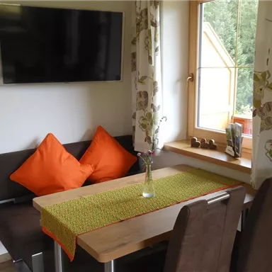 Ein gemütlicher Essbereich mit einem Holztisch und Stühlen. Die Wand ist mit einem Fernseher ausgestattet, und es gibt einen großen Fenster mit Vorhängen.