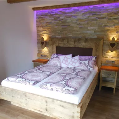 Ein gemütliches Schlafzimmer mit einem großen Bett und einem rustikalen Holzrahmen. Die Wände sind mit Stein gestaltet und sanftes Licht in Lila sorgt für eine entspannte Atmosphäre.