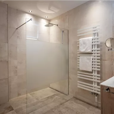 A modern bathroom with a walk-in shower and elegant tiles.  
Towels hang on a towel holder and a sink with a wooden base cabinet is visible.