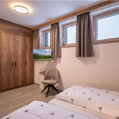 A modern bedroom with two single beds and a large wooden wardrobe. The windows are furnished with curtains and a TV adds to the amenities.