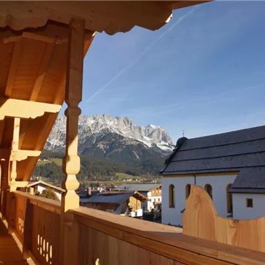 Ein Balkon mit Holzverkleidung und Blick auf eine malerische Kirch und majestätische Berge. Der Himmel ist klar und die Architektur ist traditionell.