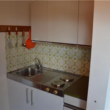 Eine kleine Küche mit einem Waschbecken und einfachen Küchenschränken. Die Wände sind mit einem Vintage-Muster dekoriert.