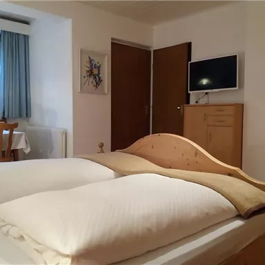A cozy bedroom with a double bed and white pillows. In the background, there is a table, a television, and a window with curtains.