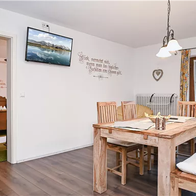 Ein helles Esszimmer mit einem großen Holztisch und Stühlen. An der Wand hängt ein Fernseher, und es gibt eine Tür zum Außenbereich.