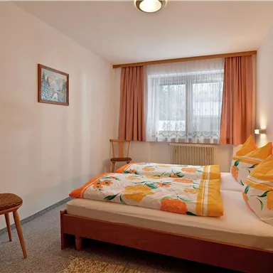 Ein gemütliches Schlafzimmer mit zwei Betten und bunten Bettwäsche. Der Raum hat ein Fenster mit Vorhängen und einen Stuhl in der Ecke.