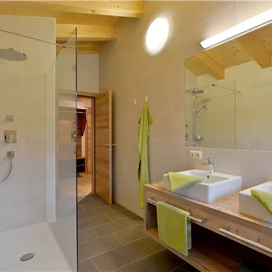 A modern bathroom with a large shower and two sinks. The walls are bright, and the decor is accented in neutral colors with green towels.