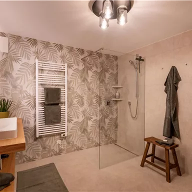 A modern bathroom with a walk-in shower and stylish plants. The walls are adorned with a tropical leaf pattern, and there is a practical wooden chair.