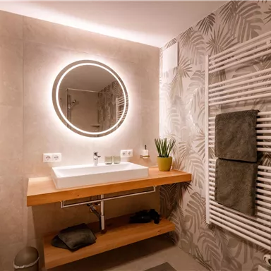 A modern bathroom with a round mirror lamp and an elegant wooden washbasin. The walls are decorated with a floral pattern, and there is a towel radiator.