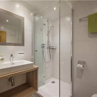 Ein modernes Badezimmer mit einer Dusche und einem Waschbecken. Die Handtücher sind grün, und die Wände sind hell gefliest.