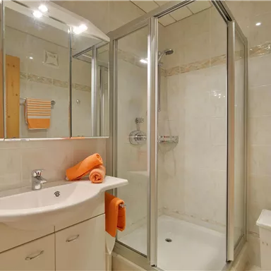 Ein modernes Badezimmer mit einer Duschkabine aus Glas und weißen Fliesen. Handtücher in Orange verleihen dem Raum einen warmen Akzent.