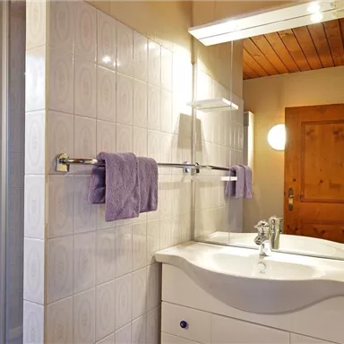 Ein modernes Badezimmer mit einem hellen Waschtisch und Handtüchern in Violett. Die Wände sind gefliest und das Licht schafft eine angenehme Atmosphäre.