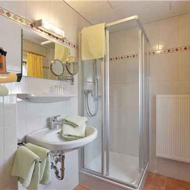 Ein helles Badezimmer mit einer Duschkabine, einem Waschbecken und Handtüchern in sanften Grüntönen. Große Fenster sorgen für natürliches Licht und eine freundliche Atmosphäre.