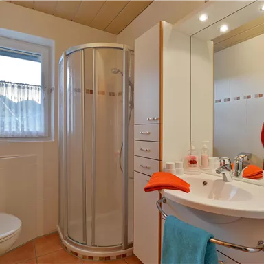 Ein modernes Badezimmer mit einer Dusche, einem Waschbecken und hellen Wänden. An den Wänden sind Handtücher in orange und blau dekoriert.