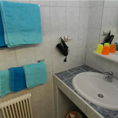 Ein Badezimmer mit blauen Handtüchern und einem weißen Waschbecken. Auf der Ablage stehen farbige Becher und ein Haartrockner ist an der Wand befestigt.
