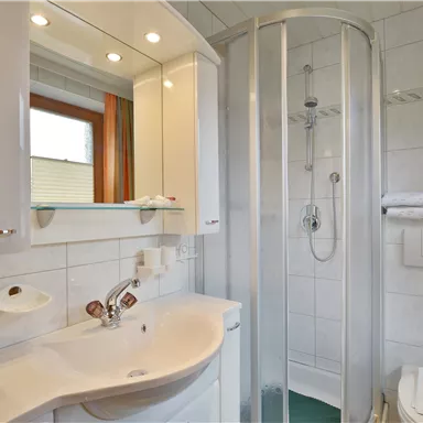 A modern bathroom with a shower and a sink. The walls are tiled, and there are large windows for natural light.