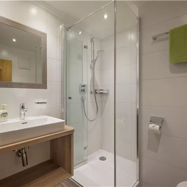 Ein modernes Badezimmer mit einer Dusche und einem Waschbecken. Die Handtücher sind grün, und die Wände sind hell gefliest.