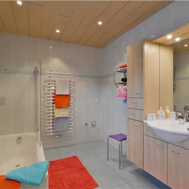 Ein helles Badezimmer mit einer Badewanne und einem modernen Waschbecken. Bunte Handtücher und ein Stuhl sorgen für Gemütlichkeit.