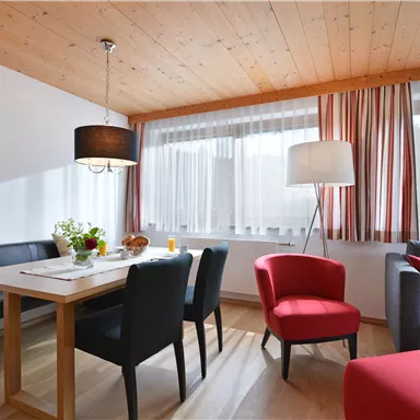 Ein gemütliches Wohnzimmer mit einem Esstisch, Stühlen und einer modernen Lampe. Große Fenster lassen viel Licht herein und schaffen eine einladende Atmosphäre.