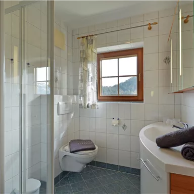 Ein modernes Badezimmer mit einer Dusche und einem Fenster. Es hat helle Fliesen und Holzdetails, sowie Handtücher auf einem Regal.