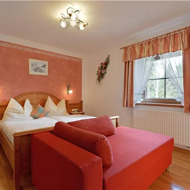 Ein gemütliches Schlafzimmer mit einem Doppelbett und einer roten Couch. Die Wände sind in sanften Farben gehalten, mit einem Fenster, das Tageslicht hereinlässt.