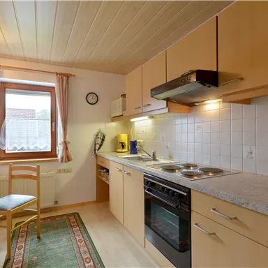 Eine helle Küche mit Holzschränken und modernen Geräten. Ein Tisch mit einem Stuhl steht in der Ecke, und das Fenster lässt viel Licht herein.