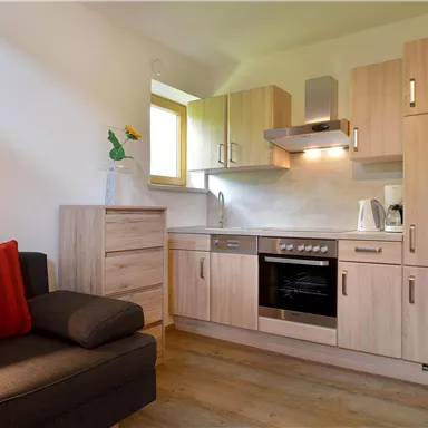 Eine moderne Küche mit Holzschränken und einem Herd. Neben der Küche befindet sich ein gemütliches Sofa mit roten Kissen.