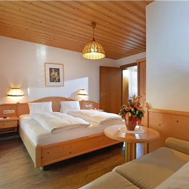 A cozy hotel room with two beds and a bright wooden ceiling. In front of the window are a table with floral decoration and a comfortable sofa.