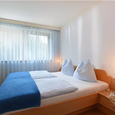 Ein helles Schlafzimmer mit einem Doppelbett und blauen Decken. Große Fenster sorgen für viel Tageslicht und eine gemütliche Atmosphäre.