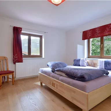 A cozy bedroom with a wooden bed and several pillows. Two windows let in plenty of light and provide a view outside.